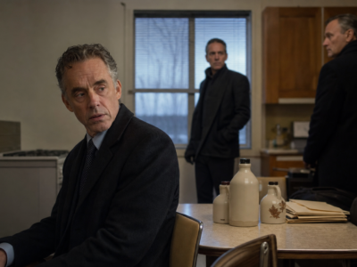 Dr. Jordan B. Peterson sits in a plain safe-house kitchen beside maple syrup jugs and folders while two plainclothes agents stand nearby.
