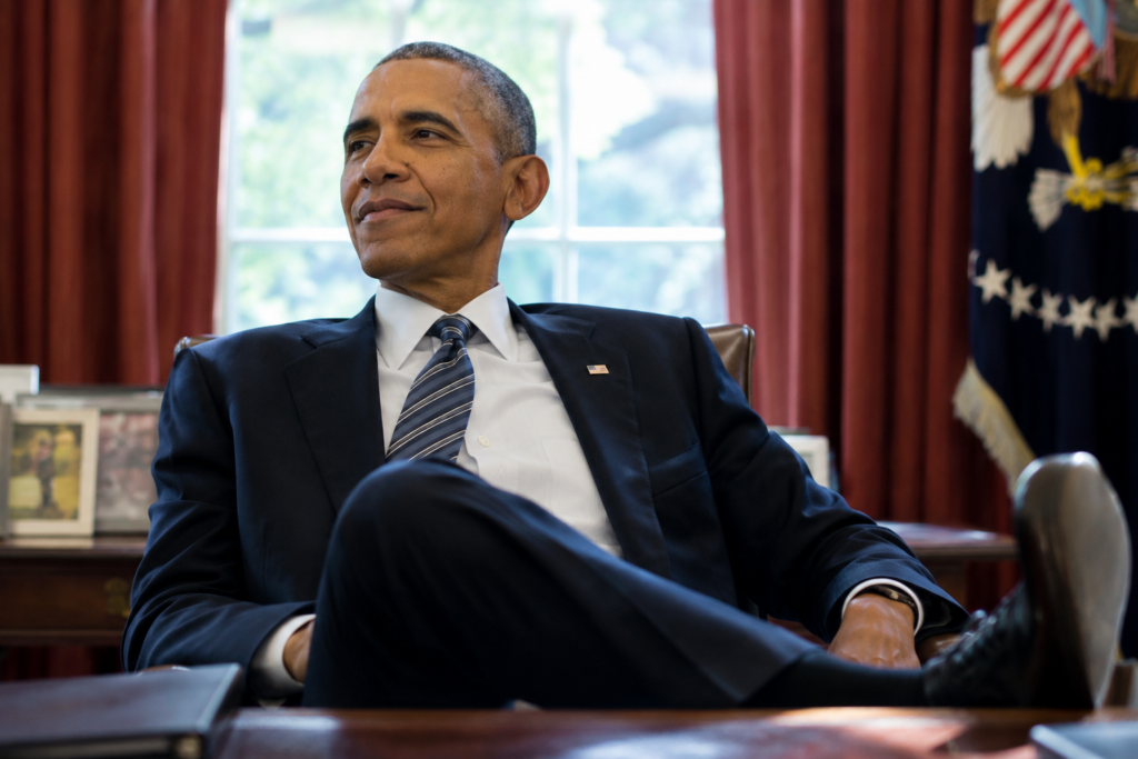Barack Obama seated in the Oval Office in front of red curtains and a presidential flag