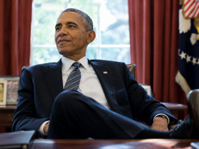 Barack Obama seated in the Oval Office in front of red curtains and a presidential flag
