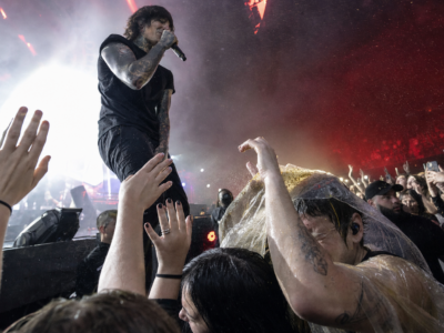 A tattooed rock singer at the edge of an arena stage as front-row fans recoil under dramatic concert lighting.