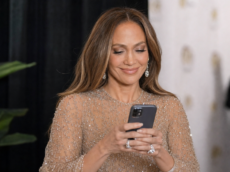 Jennifer Lopez smiles while looking at a smartphone backstage at a celebrity event.