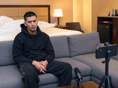 Sneako sitting on a gray hotel-room couch with a phone and microphone set up nearby.