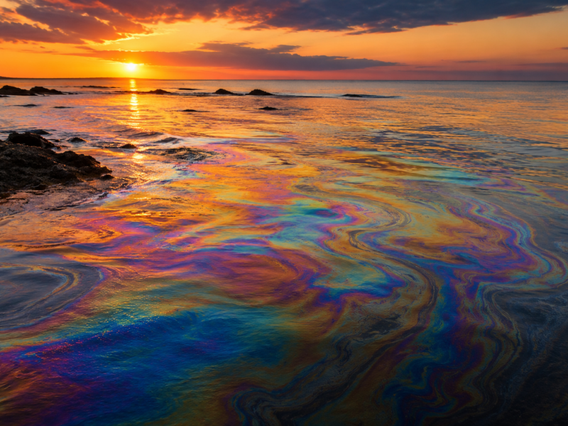 Iridescent oil slick glowing across ocean water at sunset for a satirical opinion column defending oil spills