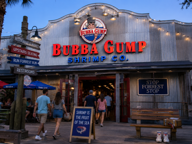 A Bubba Gump Shrimp Co. restaurant storefront with red signage, boardwalk seating, and customers walking inside.
