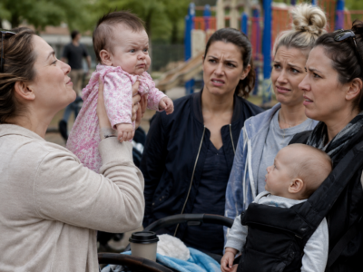 A mother holding a stern-looking baby while other mothers look confused at a playground.