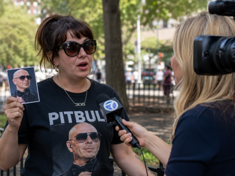 A woman wearing a Pitbull shirt holding a photo of the musician during a news interview.