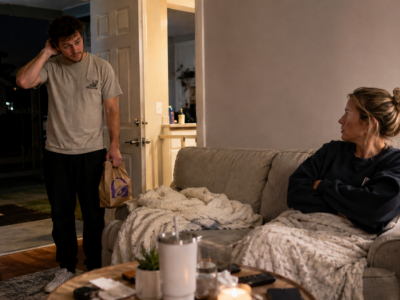 A nervous boyfriend returning home late with a fast food bag while his girlfriend waits on the couch.