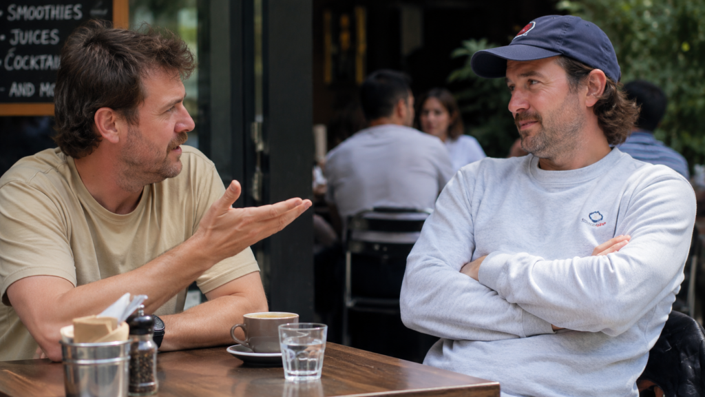 Two Australian men talking at an outdoor cafe while one cautiously explains how things are not too bad.