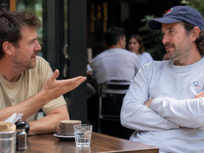 Two Australian men talking at an outdoor cafe while one cautiously explains how things are not too bad.