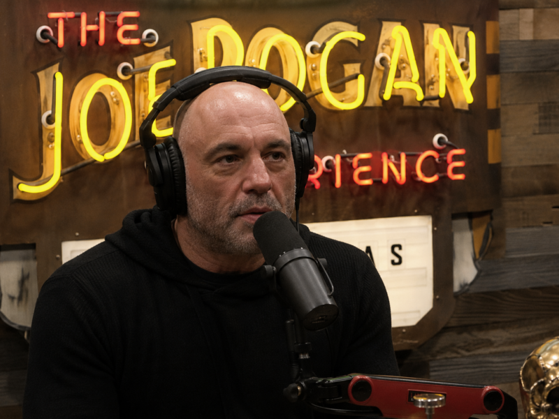 Joe Rogan speaking into a podcast microphone in front of The Joe Rogan Experience sign