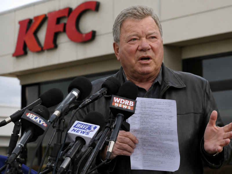 William Shatner speaking at microphones outside a KFC while holding a handwritten recipe page