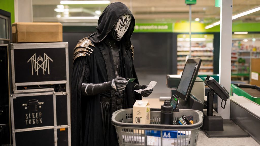 Vessel from Sleep Token stands in full ritual costume at a supermarket self-checkout with oat milk, batteries, and merch cases nearby.