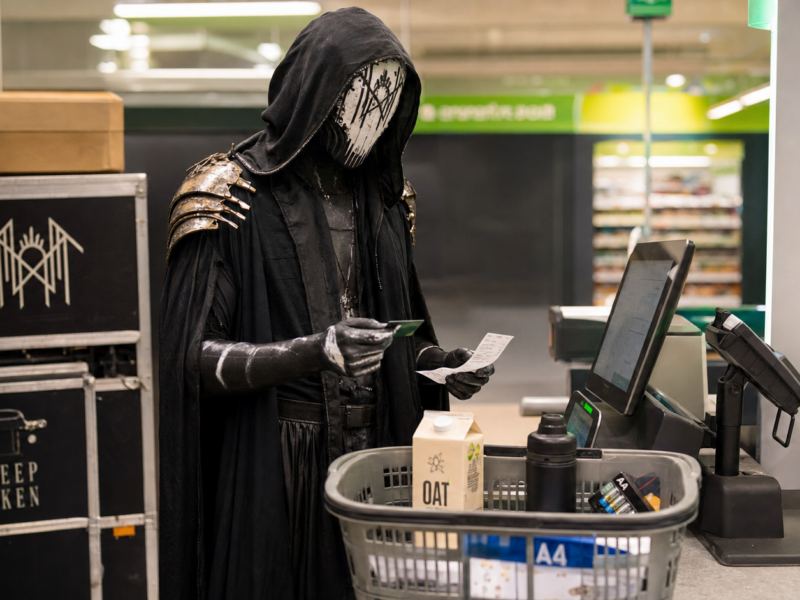 Vessel from Sleep Token stands in full ritual costume at a supermarket self-checkout with oat milk, batteries, and merch cases nearby.