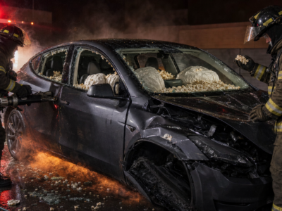 Firefighters stand beside a crashed electric car with closed doors, deployed airbags, smoke, and popcorn scattered through the cabin.