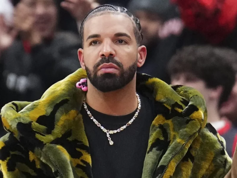 Drake sitting courtside in a green patterned coat, looking serious in a crowded arena.