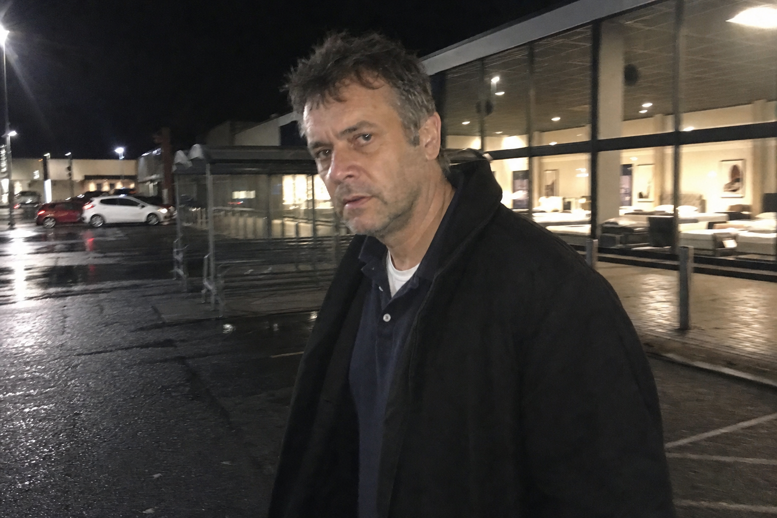 A grainy phone photo of a serious middle-aged man outside a generic British retail park at night.