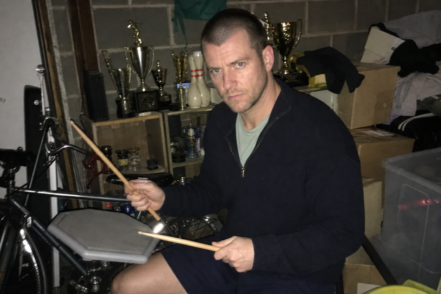 A rough phone photo of a focused man with drumsticks in a cluttered garage with bowling trophies nearby.