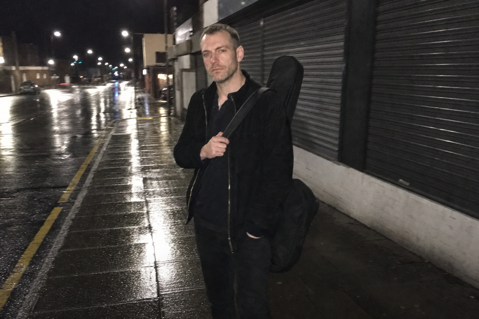 A grainy phone photo of a stylish man with a bass case outside a closed British shop on a wet evening.