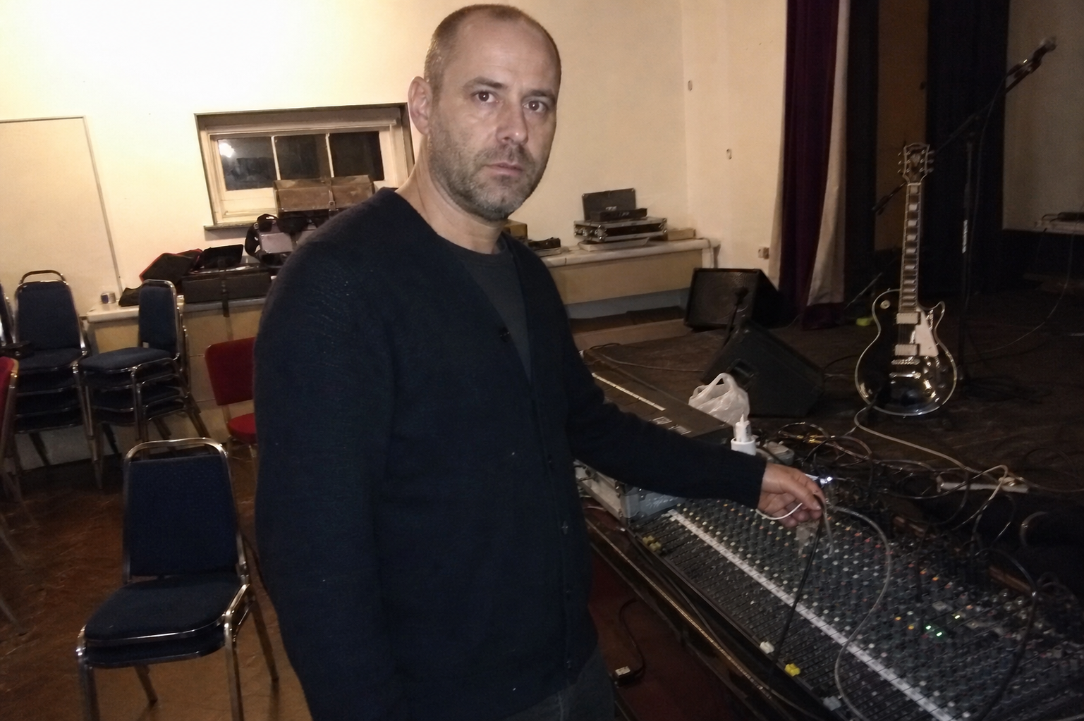 A rough phone photo of a thoughtful man beside a church hall sound desk with guitar cables nearby.