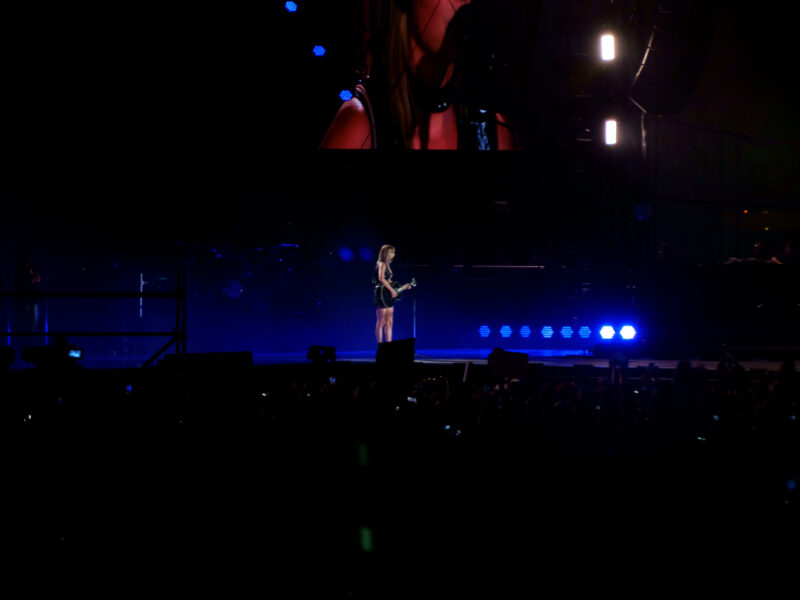 Taylor Swift performs on a dark concert stage with a large screen behind her.